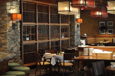 Cozy restaurant interior with stone walls and wooden wine racks. Several tables are set with white tablecloths and neatly arranged chairs. Warm lighting from wall sconces and overhead lamps creates an inviting ambiance.