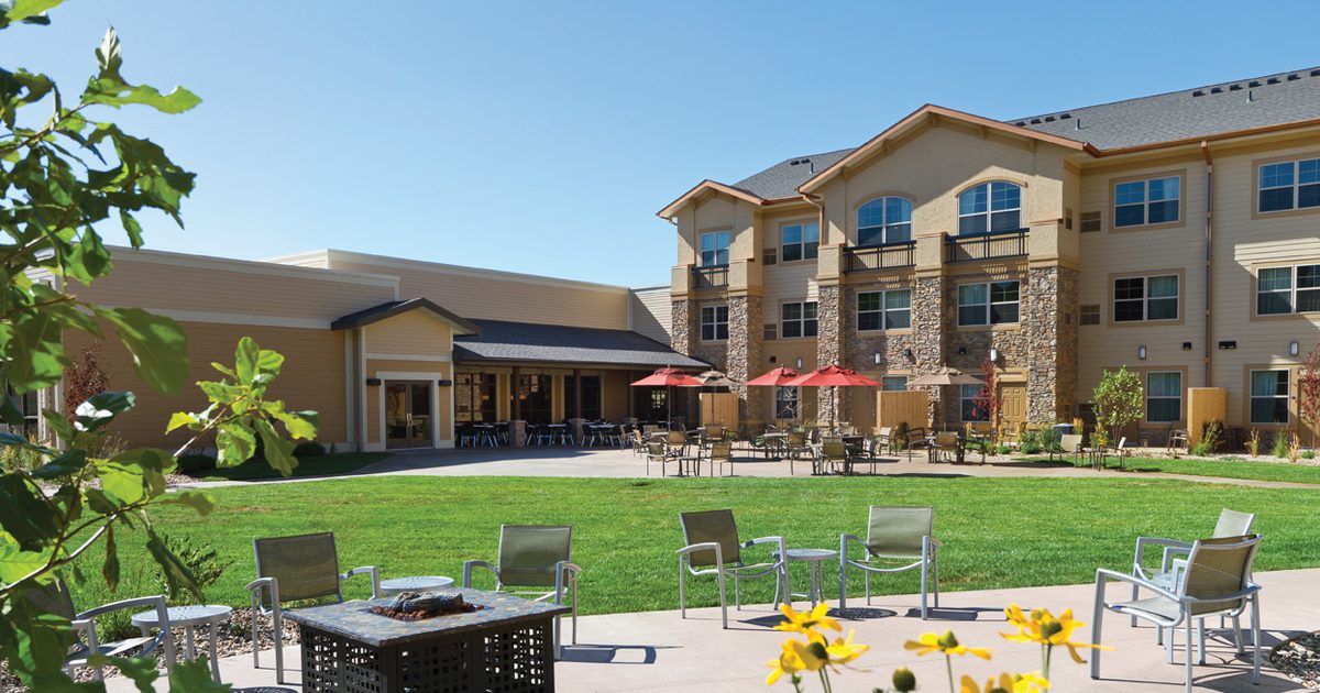 A bright courtyard with green grass and yellow flowers, surrounded by a three-story beige building. Patio chairs encircle a square fire pit, and umbrellas shade outdoor tables. Clear blue sky above.