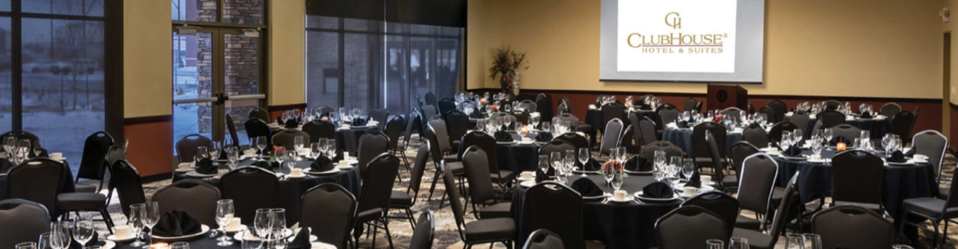 A conference room is set up for an event with round tables covered in black tablecloths. Each table is arranged with glassware, plates, and napkins. A projector screen displays the "ClubHouse Hotel & Suites" logo at the front of the room.