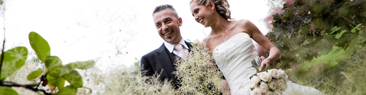 A joyful couple is standing outdoors in a lush, green setting. The bride is in a white strapless wedding dress holding a bouquet of roses, and the groom is in a black suit and tie. They are smiling and looking at each other.